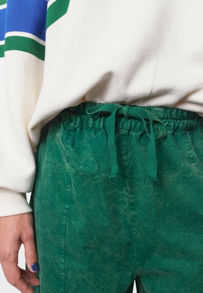 Groene, gestructureerde broek met een elastische tailleband en bijpassend trekkoord, gecombineerd met een witte trui met blauwe en groene strepen.