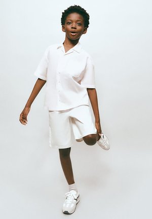 Băiat tânăr în cămașă albă și pantaloni scurți, echilibrându-se pe un picior, ținându-și glezna, pe un fundal simplu, deschis la culoare.