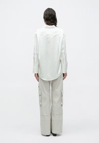 Persoană cu păr lung și închis la culoare, purtând o cămașă albă largă din satin și pantaloni cargo gri deschis, stând cu spatele, pe un fundal simplu.