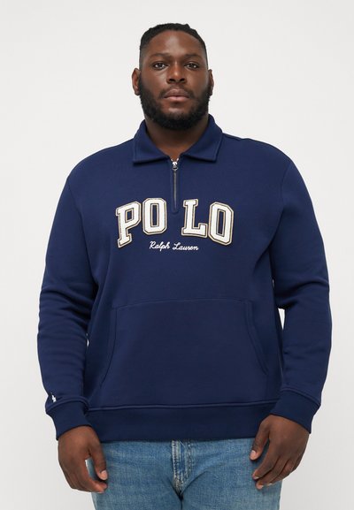 Dunkelblaues Zip-Up-Sweatshirt mit hochschließendem Kragen, versehen mit weißem, gesticktem "POLO"-Text und einer Fronttasche. Das Material wirkt weich und strukturiert.