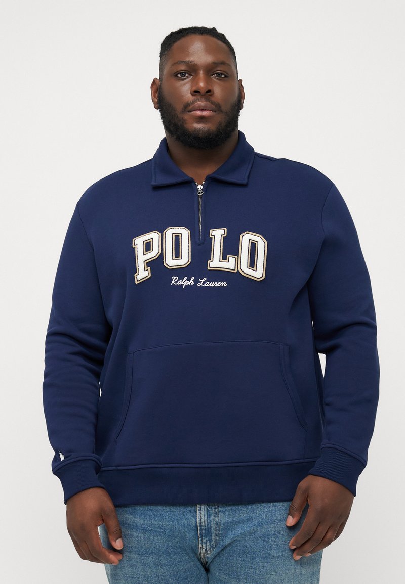 Marineblauwe rits-jas met hoge kraag, voorzien van witte geborduurde "POLO" tekst en een voorzak. Materiaal lijkt zacht en gestructureerd.