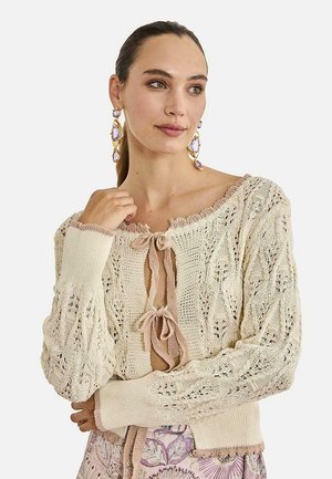 Mujer con el pelo recogido lleva un cárdigan de punto color crema con dos lazos delanteros, pendientes llamativos color lavanda y falda estampada.