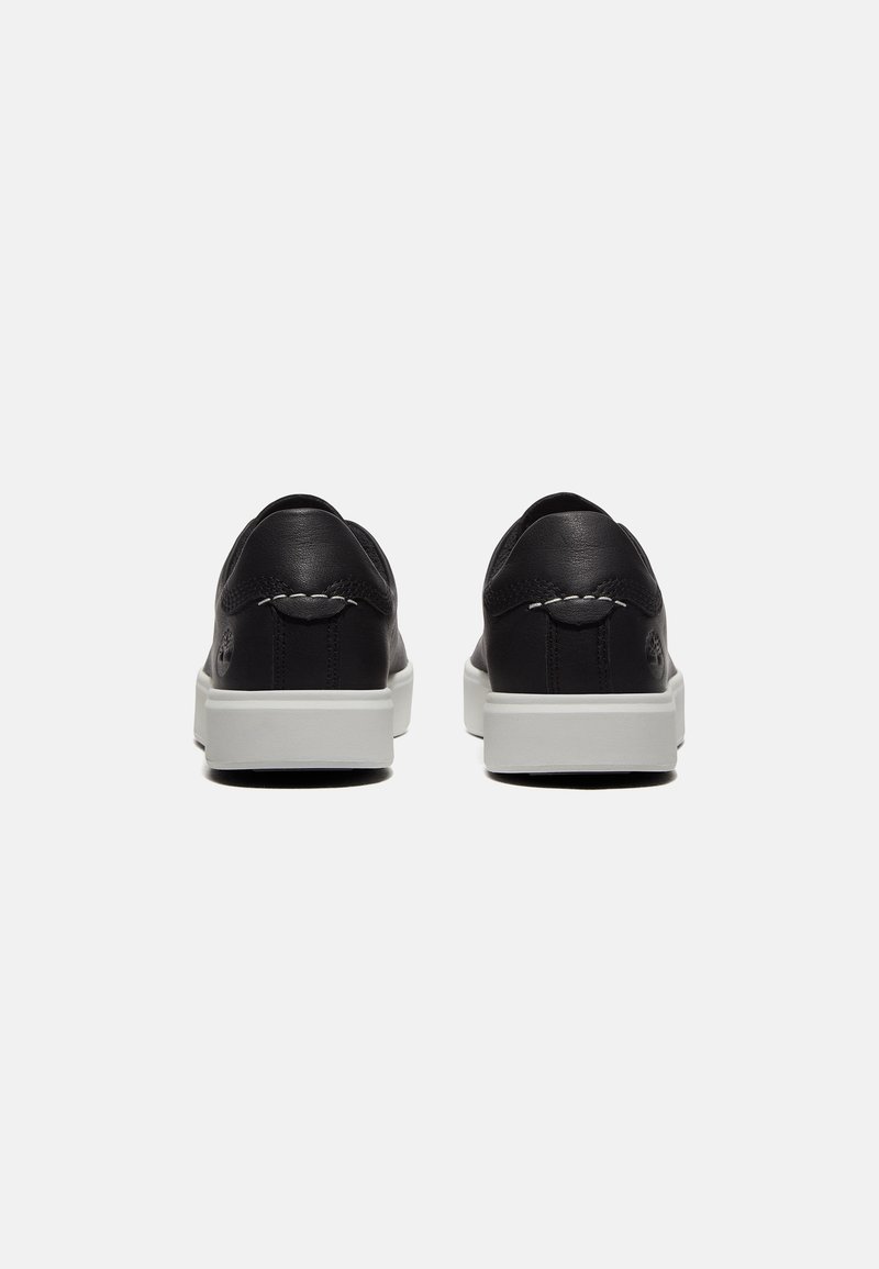 Zapatillas de cuero negras con una suela de goma blanca. Vista trasera que muestra una textura suave, costuras mínimas y una marca sutil en el talón.