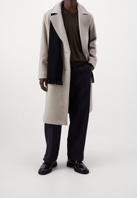 Manteau en laine beige avec grands revers, écharpe noire à franges, chemise vert olive en V, pantalon sombre et chaussures noires brillantes.