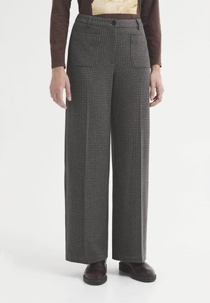 Persona con pantalones anchos de talle alto en gris y negro con estampado de pata de gallo, con bolsillos delanteros y zapatos marrón oscuro, de pie sobre fondo blanco.