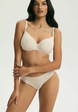 Kobieta o długich ciemnych włosach, ubrana w jasnobeżowy biustonosz i dopasowaną bieliznę, stojąca z rękami skrzyżowanymi na brzuchu.