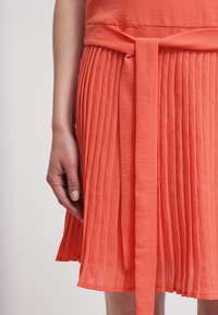 Robe corail plissée avec ceinture nouée à la taille et tissu léger. La jupe présente des plis verticaux pour la texture et le mouvement.