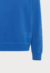 Blauwe sweatshirt met ribgebreide manchetten en zoom, voorzien van textuur borduurwerk met de tekst "ELB" en "SAND" op de mouw. Zacht materiaal, casual ontwerp.
