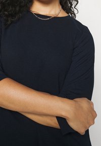 Marinblå topp med trekvartslång ärm, strukturerat tyg, rund halsringning och ett lager av silverkedjor. Modellens armar är korsade.