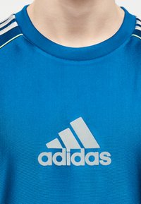 Μπλε αθλητική μπλούζα Adidas με λευκό λογότυπο και ρίγες στους ώμους, φορεμένη από άτομο, σε θέα από τον λαιμό έως το επάνω μέρος του στήθους.