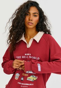 Vínově červená oversized mikina s béžovým límcem a grafickým potiskem zobrazením hrnků kávy a žlutého taxi. Text obsahuje "VÍTÁME VÁS VE SKUTEČNÉM SVĚTĚ."