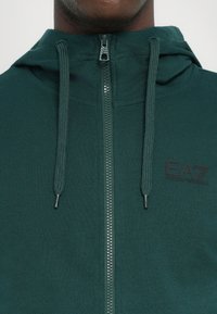 Tmavo zelená mikina na zips s textúrovaným povrchom, nastaviteľnými sťahovacími šnúrkami a logom EA7. Disponuje hladkým zipsom a tesným strihom okolo krku.