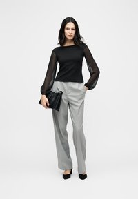 Γυναίκα με μαύρο τοπ με διάφανα μανίκια, γκρι παντελόνι φαρδύ, μαύρα ψηλοτάκουνα, κρατώντας μαύρη τσάντα, στέκεται μπροστά σε απλό λευκό φόντο.