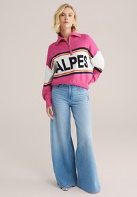 Roza pletena pulover s pol-zadrgo in bold "ALPES" napisom; v kombinaciji s širokimi svetlo modrim kavbojkam; mehka tekstura, sproščen oblikovanje.