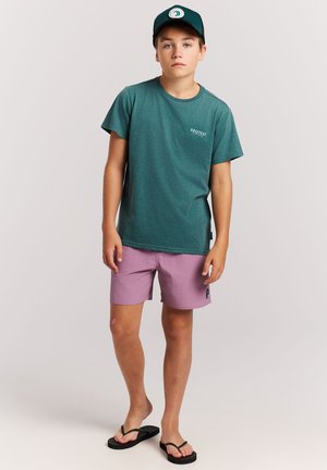 Boy wearing teal cap and t-shirt, pink shorts, and black flip-flops, standing against a plain light background.
