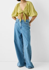 Blusa corta verde lime con maniche a palloncino e fiocco frontale, abbinata a jeans in denim blu a vita alta e gamba larga. Tessuto liscio, stile casual.