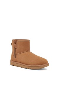 Cizme ankle din suede de culoare bej, cu o textură netedă, vârf rotunjit, fermoar pe lateral  și o talpă din cauciuc bej. Branding UGG pe patch-ul din piele.