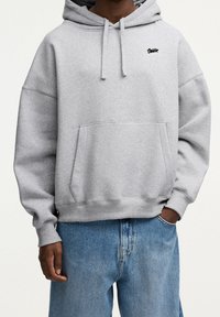 Harmaa huppari, joka on valmistettu puuvillasekoitteesta ja jossa on kengurutasku, resorireunaiset hihansuut ja pieni musta logo rinnassa. Yhdistetty sinisiin farkkuihin.