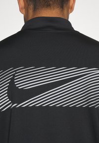 Schwarzes Polohemd mit erhöhtem Kragen, das mit kräftigen, diagonalen weißen Streifen und einem auffälligen Nike-Logo in der Mitte gestaltet ist. Strukturiertes Gewebe.