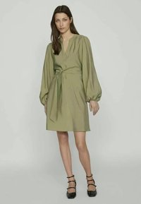 Ελαιόχρωμο κοντό φόρεμα με V-λαιμόκοψη, μακριές φουσκωτές μανίκες και ζώνη στη μέση. Απαλή υφή, στενή εφαρμογή και σχέδιο που φτάνει μέχρι το γόνατο.