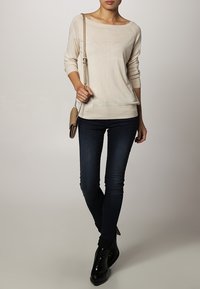 Krémový pletený svetr s dlouhým rukávem a širokým výstřihem, spárovaný s tmavými skinny džínami a lesklými černými kotníkovými botami. Drží hnědou kabelku přes rameno.
