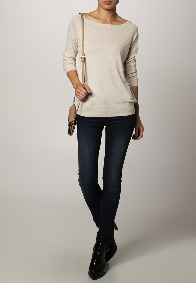 Krémový pletený svetr s dlouhým rukávem a širokým výstřihem, spárovaný s tmavými skinny džínami a lesklými černými kotníkovými botami. Drží hnědou kabelku přes rameno.