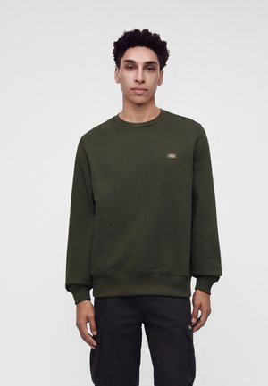 Ung mand med kort krøllet hår iført en mørkegrøn sweatshirt og sorte cargobukser, stående mod en simpel lys baggrund.
