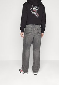 Pánská černá mikina s kapucí a grafickým potiskem na zádech, doplněná volnými, vybledlými šedými džíny. Tenisky mají červené a černé akcenty.