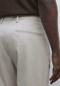 Närbild av en person som bär ljusbeige byxor och en mörkbrun skjorta, med fokus på bakfickan och höger arm.
