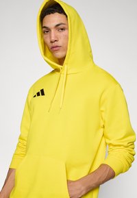 Κίτρινο φούτερ με κουκούλα από απαλό ύφασμα, με μπροστινή τσέπη, κορδόνια και μαύρο λογότυπο στην πάνω αριστερή πλευρά.