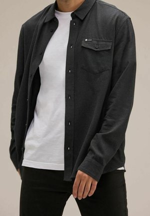 Man draagt een donkergrijs overhemd met knopen over een wit T-shirt en een zwarte broek, staand met één gedeeltelijk zichtbare hand.