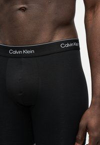 Ανδρικός κορμός που φοράει μαύρα μποξεράκια Calvin Klein, με εμφανές το λογότυπο στη ζώνη και μέρος του δεξιού χεριού, σε απλό φόντο.