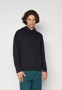 Felpa nera realizzata in morbido misto cotone; presenta una tasca frontale, cappuccio con coulisse e polsini a coste, abbinata a pantaloni verdi.