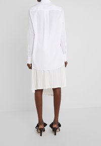 Chemise blanche à manches longues avec un ourlet incurvé, associée à une jupe plissée blanche avec fines rayures verticales, et des sandales noires à talons.