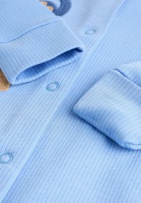 Indumento lungo a maniche ribbed di colore azzurro chiaro con due bottoni di chiusura e polsini ripiegati. Presenta un tessuto texturizzato e sottili linee verticali.