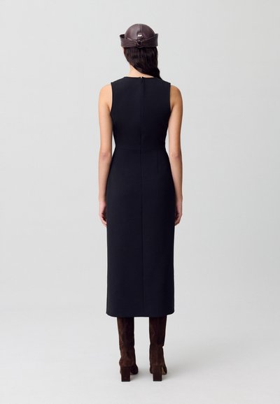 Vestido negro sin mangas con una silueta ajustada, a la altura de la rodilla, con cierre de cremallera en la espalda. Combinado con botas de ante marrón oscuro hasta la rodilla.