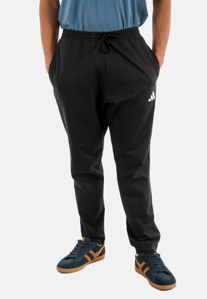Pantalon de sport noir avec une taille élastique, des poches latérales et un accent logo adidas blanc. Portés avec des baskets bleues présentant des rayures marron.
