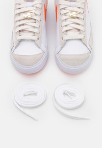 Un paio di sneaker bianche e beige con accenti arancioni, charm per lacci Nike dorati e due lacci bianchi avvolti separatamente sotto le scarpe.