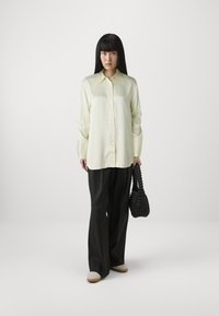 Camicia button-up gialla chiara con colletto a punta, abbinata a pantaloni neri a gamba larga e una borsa nera strutturata. Footwear casual completa il look.