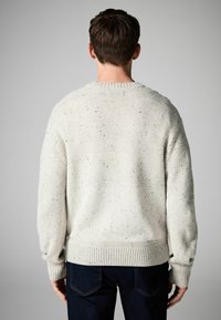 White knit sweater with a textured pattern and speckled accents, featuring ribbed cuffs and hem. Worn over dark jeans.