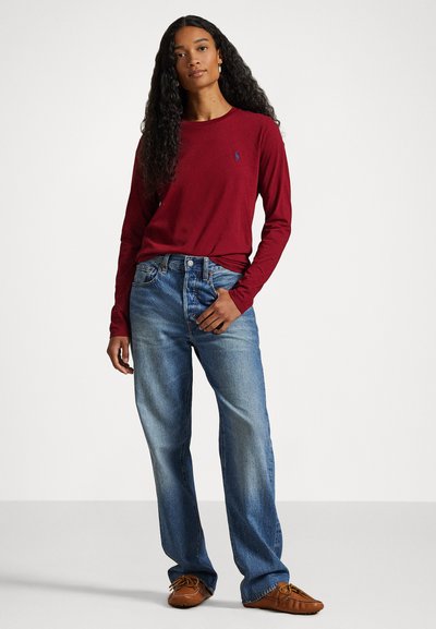 Burgundska majica s dolgimi rokavi z majhnim logotipom in svetloblue jeans hlače ravnega kroja, nosene z rjavimi mokasini.