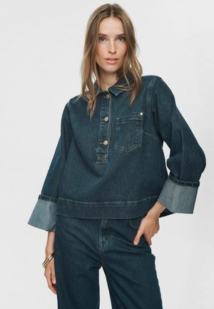 Junge Frau trägt ein dunkelblaues Denim-Hemd mit hochgekrempelten Ärmeln und passende Jeans. Sie steht mit der Hand auf der Hüfte vor einem schlichten Hintergrund.