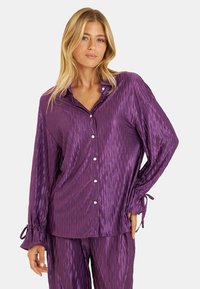 Camisa de satén púrpura con botones, con un patrón texturizado de diamantes y mangas fruncidas, que presenta botones plateados y lazos en los puños.