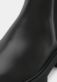 Bota de tornozelo em couro preto com textura suave, bico arredondado e sola robusta. Possui costura mínima ao longo da borda.