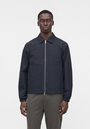 Homme portant une veste zippée bleu marine foncé avec col et poignets élastiques, associée à un pantalon gris, debout devant un fond blanc uni.