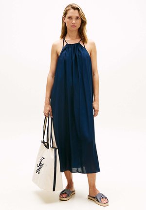 Kvinde stående iført en ærmeløs marineblå halterneck-kjole, stribede espadrille-sandaler og holder en stor hvid tote taske med sorte håndtag.