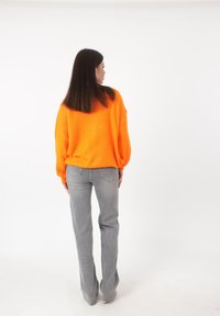 Frau mit langen dunklen Haaren, die einen orangefarbenen Pullover und graue Jeans trägt, stehend und von hinten gegen einen schlichten weißen Hintergrund gerichtet.