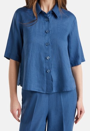 Femme portant une chemise bleue à manches courtes boutonnée et un pantalon bleu assorti, visible du cou jusqu'au milieu des cuisses sur un fond blanc.