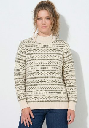 Woman with wavy hair in a high ponytail wearing a cream sweater with green geometric patterns and blue jeans against a plain background.