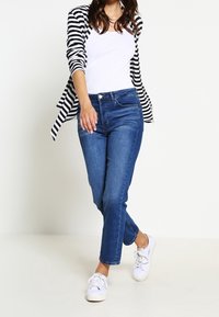 Mujer caminando en jeans azules, zapatillas blancas, camiseta blanca sin mangas y una camisa de rayas en blanco y negro sobre los hombros.